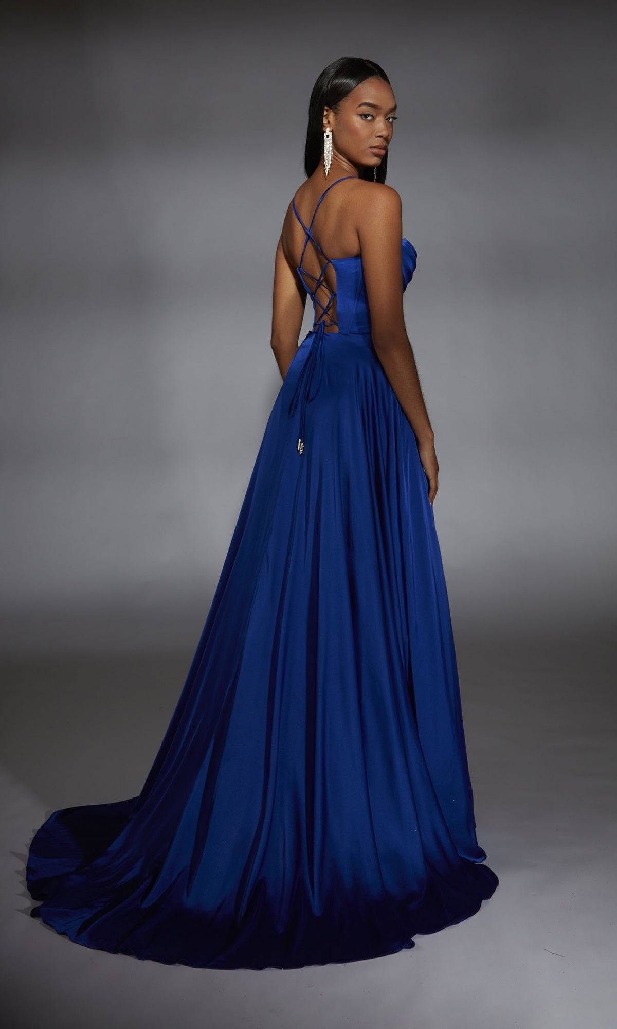 A woman wears the Alyce 61883 Long Sweetheart A-Line Prom Dress in royal blue, showcasing a sweetheart corset bodice, crisscross straps, and a lace-up open back, posed against a plain gray background.